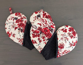 Puede incluir: Tres manoplas acolchadas en forma de corazón. Las manoplas presentan un estampado floral crema con flores rojas y detalles de lunares negros. Cada manopla tiene un lazo para colgar.