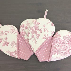 May include: Three heart-shaped oven mitts with a floral pattern in pink and white. The mitts have a loop for hanging and are made of quilted fabric. The design features a mix of floral and polka dot patterns.