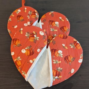 May include: Three round quilted pot holders featuring a burnt orange fabric with a repeating pattern of pumpkins, green leaves, and white flowers. Each pot holder has white fabric trim and a light blue interior lining with a subtle white snowflake print.