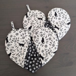May include: Three heart-shaped pot holders with a black and white cat and floral pattern. The pot holders have a loop for hanging and are made of fabric. The bottom section of the pot holders has a black and white paw print pattern.