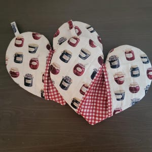 May include: Two heart-shaped pot holders with a red and white checkered pattern on the back. The front of the pot holders features a white background with a repeated pattern of jars of jam in various colors.