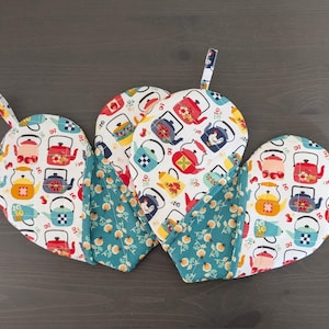 May include: A set of three heart-shaped oven mitts. The mitts feature a white background with colorful teapots. The center of the mitts are a teal floral pattern. Each mitt has a small loop for hanging.