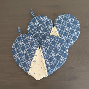 May include: Four heart-shaped oven mitts with a blue and white floral pattern. The mitts have a quilted design with a white grid and small blue flowers. A cream-colored fabric with blue floral accents is visible between the mitts.