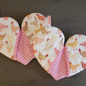 May include: Two heart-shaped pink and white pot holders with a chicken print. The pot holders have a pink and white checkered pattern on the back.