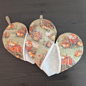 May include: A double oven mitt featuring a sage green fabric with a repeating autumn harvest print of orange pumpkins, red wagons, and sunflowers. The mitt has a quilted texture, white fabric trim, and two fabric loops for hanging storage.