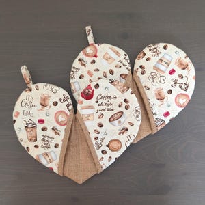 May include: Three heart-shaped oven mitts with a coffee-themed pattern. The mitts feature a beige background with coffee beans, cups, and text like "It's Coffee Time" and "Coffee is always a good idea." The edges are trimmed with a brown fabric.