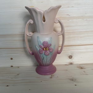 May include: A vintage pink ceramic vase with a floral design. The vase has two handles and a flared top. A raised flower with a pink center and yellow accents is centered on the front. The vase is on a light wood surface.