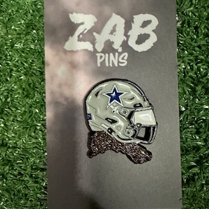 May include: A silver and blue enamel pin featuring a football helmet with a star on the side. The pin is on a gray card with the text "ZAB PINS" printed in white.
