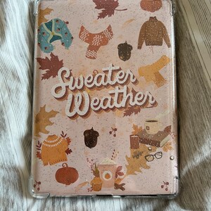 May include: A pink glitter phone case with a fall-themed design. The case features a variety of autumnal elements, including leaves, pumpkins, sweaters, and coffee cups. The text "Sweater Weather" is printed in a stylized font.