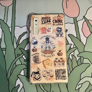 May include: A clear phone case with a beige background and various colorful stickers. The stickers feature coffee, books, ghosts, and spooky sayings like "More Espresso Less Depresso" and "I Like My Books Spicy".