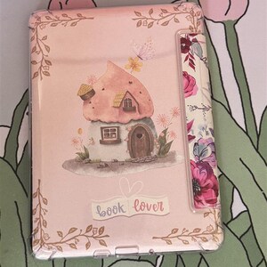 以下が含まれることがあります： ピンクの花柄とキノコの家が描かれたピンクのブックカバー。カバーには「book lover」の文字が印刷されています。