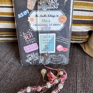 Puede incluir: Un lector electrónico Kindle negro con una funda protectora transparente. La funda está decorada con pegatinas, incluyendo una taza de café, una flor, una cara sonriente y un murciélago. El texto en la funda dice "This kindle belongs to Maria I'M READING, GO AWAY! DUH!" El Kindle está unido a una cadena de cuentas rosa y blanca con un dije de flor rosa.
