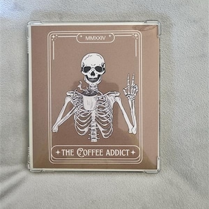 Puede incluir: Una tarjeta marrón con un esqueleto blanco sosteniendo una taza de café. La tarjeta tiene el texto "MMXXIV" y "THE COFFEE ADDICT".
