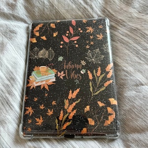 May include: A black and clear phone case with a glittery autumn-themed design. The design features leaves, branches, and the text "Autumn Vibes".
