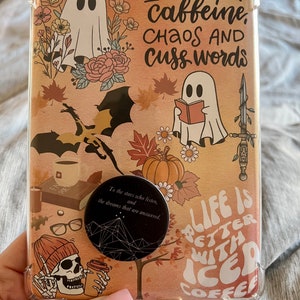 May include: A clear phone case with a colorful design featuring ghosts, dragons, pumpkins, and fall leaves. The case has a pop socket with a black and white design. The text on the case reads "I run on caffeine, chaos and cuss words" and "Life is better with iced coffee".