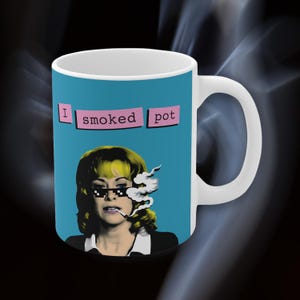 Pode incluir: Caneca de cerâmica branca com fundo azul apresentando uma imagem em estilo pop art de uma mulher fumando. O texto "I smoked pot" está acima da imagem em caixas rosa. A caneca tem uma alça branca.