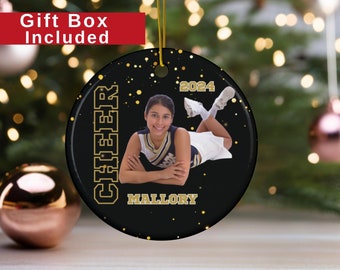 May include: A round, black and gold ceramic ornament with a photo of a cheerleader. The word "CHEER" is printed vertically, and "MALLORY" is printed below the photo. The year "2024" is printed at the top. A gold string is attached for hanging.