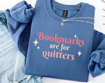 May include: A blue crew neck sweatshirt with the text "Bookmarks are for quitters" in pink and yellow. The sweatshirt is paired with blue jeans and a silver necklace. The design includes yellow starburst graphics.