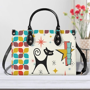 Könnte beinhalten: Cremefarbene Handtasche mit einer schwarzen Katzenillustration und einem Retro-Starburst-Design. Die Tasche hat einen schwarzen Griff und Riemen sowie ein farbenfrohes geometrisches Muster an den Seiten. Die Tasche ist rechteckig.