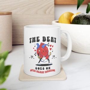 May include: White ceramic mug featuring a cartoon heart character with a stethoscope, musical notes, and hearts. The text on the mug reads "THE BEAT GOES ON OPEN HEART WARRIOR" with the years "20" and "24" on either side of the heart.