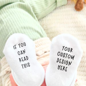 May include: White baby socks with black text that reads "IF YOU CAN READ THIS" on one sock and "YOUR CUSTOM DESIGN HERE" on the other. The socks are on a baby's feet, with a light green outfit and cream-colored blanket in the background.