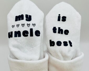 May include: A pair of white baby socks with black text. One sock says "my uncle" with hearts and the other sock says "is the best".