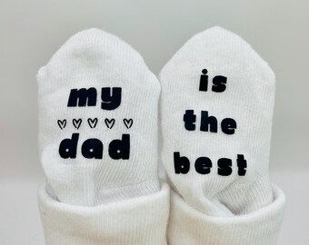 May include: A pair of white baby socks with black text. One sock says "my dad" with hearts and the other sock says "is the best".