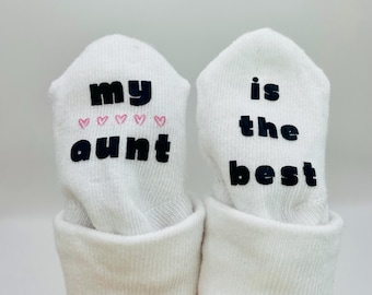 May include: A pair of white baby socks with black text. One sock says "my aunt" with pink hearts. The other sock says "is the best".