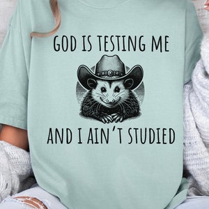 May include: A light blue t-shirt with a black and white graphic of a possum wearing a cowboy hat. The text on the shirt reads "God is testing me and I ain't studied."