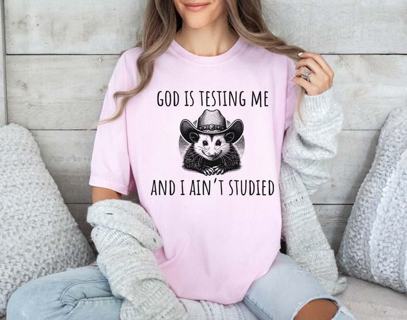 May include: Pink t-shirt with a black and white illustration of a possum wearing a cowboy hat. The text "God is testing me and I ain't studied" is printed on the shirt.