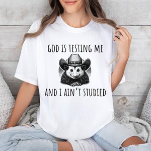 May include: White t-shirt with a black and white illustration of a possum wearing a cowboy hat. The text "God is testing me and I ain't studied" is printed on the shirt.