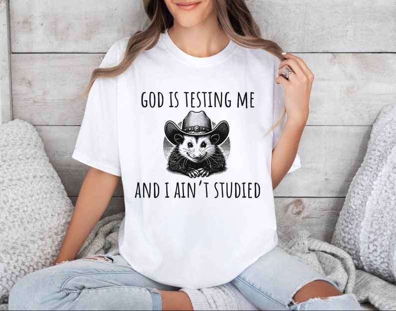 May include: White t-shirt with a black and white illustration of a possum wearing a cowboy hat. The text "God is testing me and I ain't studied" is printed on the shirt.