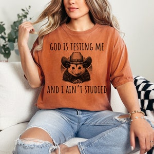 May include: A woman wearing a burnt orange t-shirt with a black graphic of a possum wearing a cowboy hat. The text on the shirt reads "God is testing me and I ain't studied".