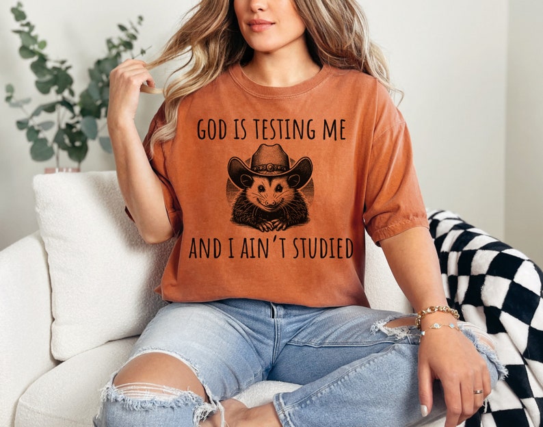 May include: A woman wearing a burnt orange t-shirt with a black graphic of a possum wearing a cowboy hat. The text on the shirt reads "God is testing me and I ain't studied".