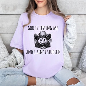 May include: A lavender t-shirt with a graphic of a possum wearing a cowboy hat. The text on the shirt reads "God is testing me and I ain't studied."