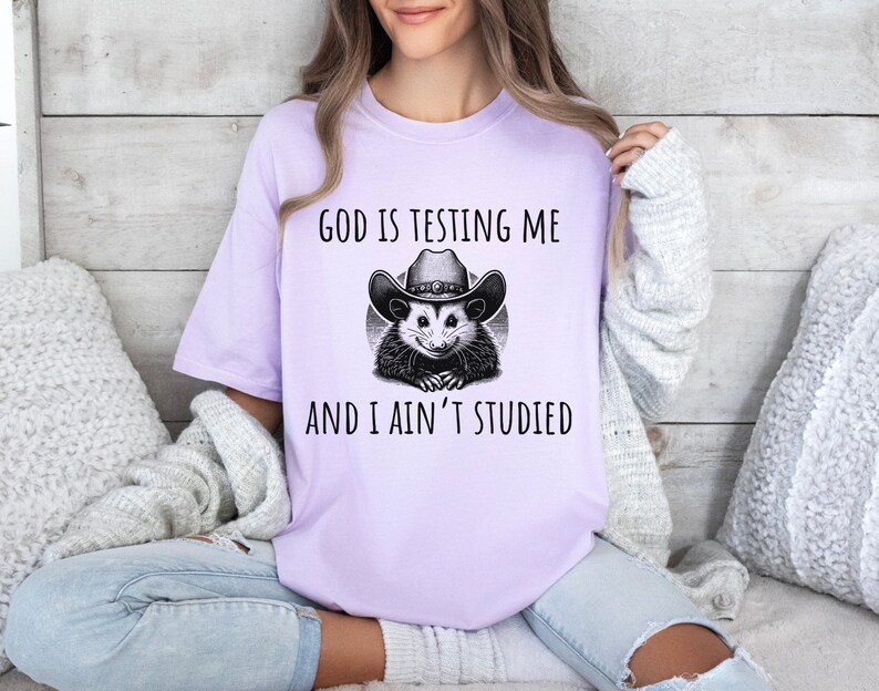 May include: A lavender t-shirt with a graphic of a possum wearing a cowboy hat. The text on the shirt reads "God is testing me and I ain't studied."