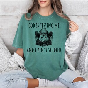 May include: A green t-shirt with a black and white graphic of an opossum wearing a cowboy hat. The text on the shirt reads "God is testing me and I ain't studied."