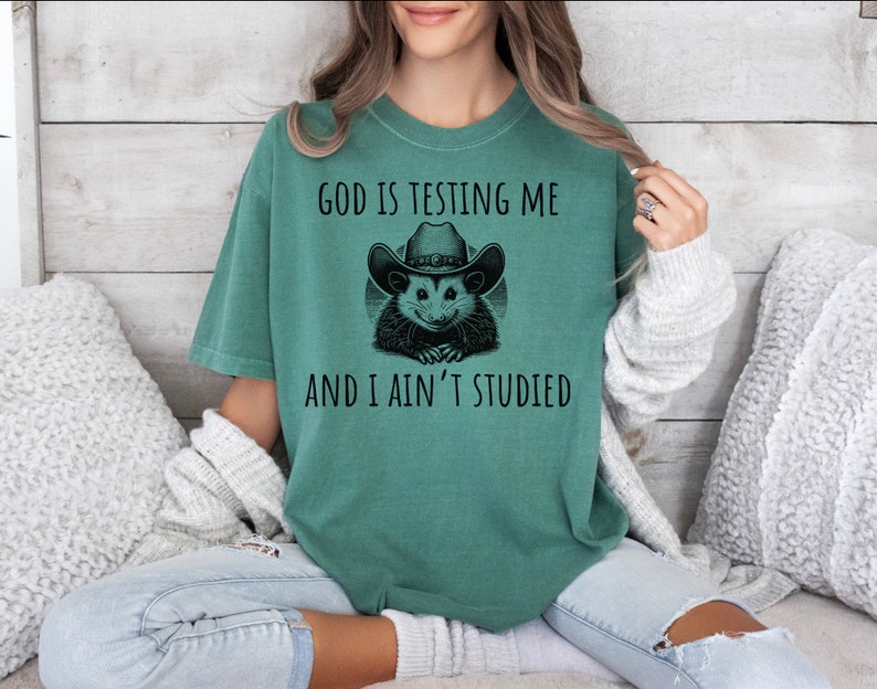 May include: A green t-shirt with a black and white graphic of an opossum wearing a cowboy hat. The text on the shirt reads "God is testing me and I ain't studied."