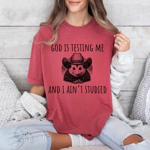 May include: Red t-shirt with a black and white illustration of an opossum wearing a cowboy hat. The text "God is testing me and I ain't studied" is printed on the shirt.