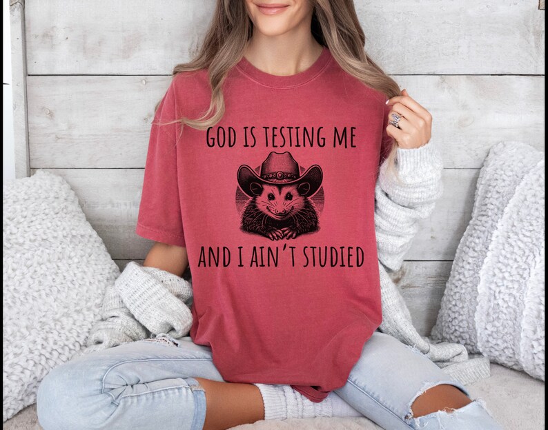 May include: Red t-shirt with a black and white illustration of an opossum wearing a cowboy hat. The text "God is testing me and I ain't studied" is printed on the shirt.