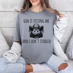 May include: A gray t-shirt with a black and white illustration of an opossum wearing a cowboy hat. The text "God is testing me and I ain't studied" is printed on the shirt.