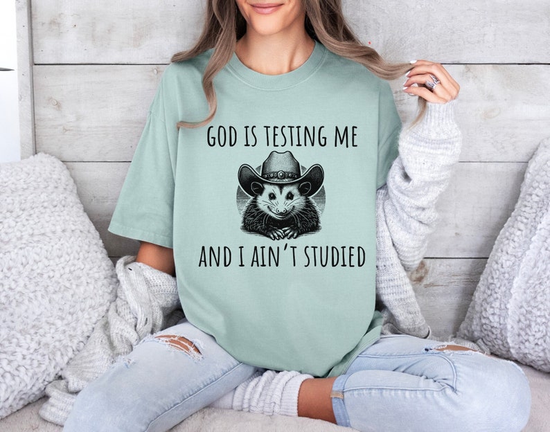 May include: A light blue t-shirt with a black and white graphic of a possum wearing a cowboy hat. The text on the shirt reads "God is testing me and I ain't studied."