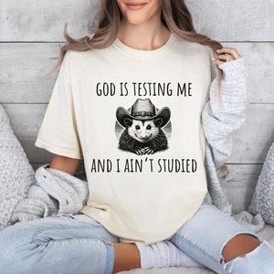 May include: A white t-shirt with a black and white illustration of an opossum wearing a cowboy hat. The text "God is testing me and I ain't studied" is printed on the shirt.
