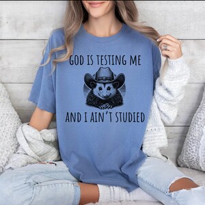 May include: A blue t-shirt with a graphic of a possum wearing a cowboy hat. The text on the shirt reads "God is testing me and I ain't studied."