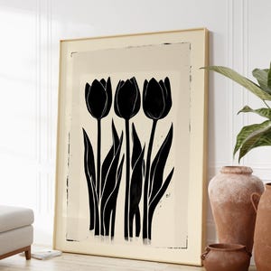 May include: A framed art print featuring a minimalist design of three black tulip flowers with long stems and leaves against a cream-colored background. The artwork is in a gold frame and leans against a white wall.