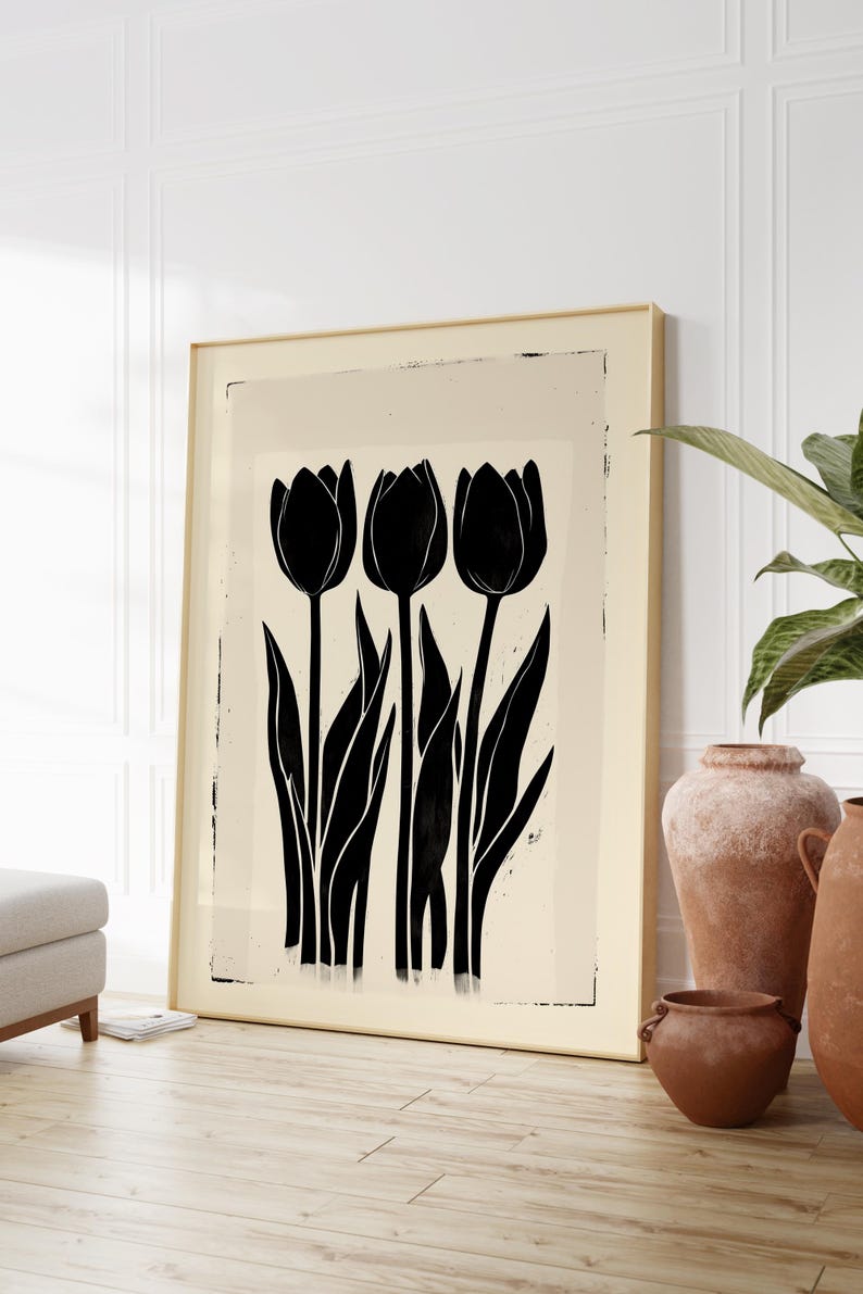 May include: A framed art print featuring a minimalist design of three black tulip flowers with long stems and leaves against a cream-colored background. The artwork is in a gold frame and leans against a white wall.