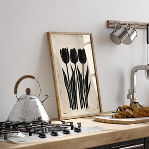 May include: A framed art print featuring a black and white illustration of three tulips. The artwork is displayed on a kitchen counter next to a stovetop, a kettle, and a cutting board with sliced bread. The frame is made of wood.