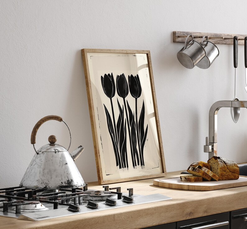 May include: A framed art print featuring a black and white illustration of three tulips. The artwork is displayed on a kitchen counter next to a stovetop, a kettle, and a cutting board with sliced bread. The frame is made of wood.
