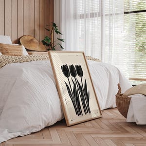 May include: A framed art print featuring a black and white tulip design. The artwork is displayed in a light-colored wooden frame and leans against a bed with white bedding. The print has a minimalist aesthetic.