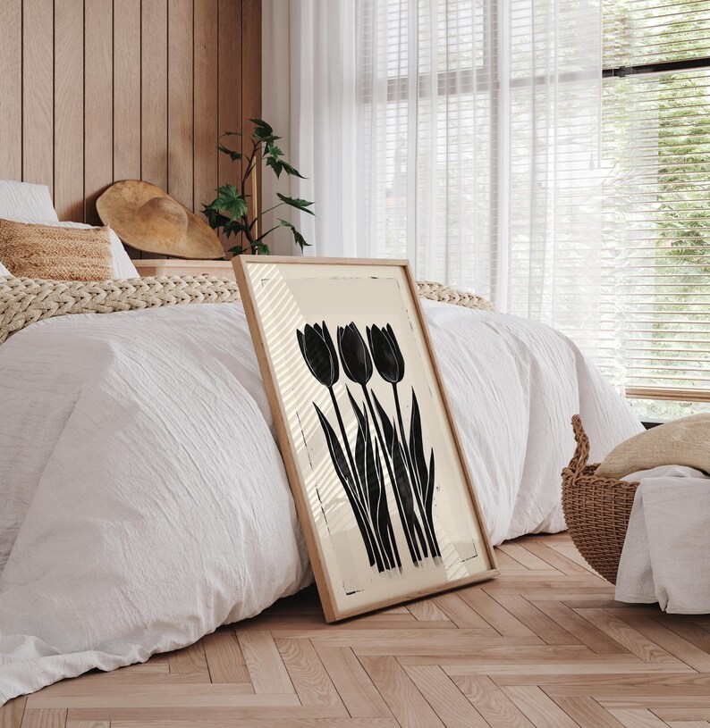 May include: A framed art print featuring a black and white tulip design. The artwork is displayed in a light-colored wooden frame and leans against a bed with white bedding. The print has a minimalist aesthetic.